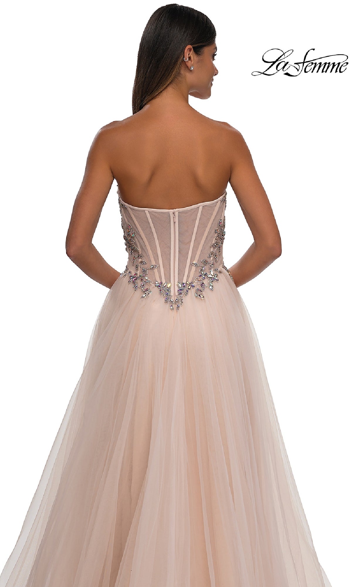 A woman stands facing away in the La Femme 32862 Beaded-Corset Strapless Prom Gown featuring a fitted, semi-sheer beaded bodice with floral embellishments and a long a-line skirt.